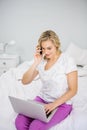 Woman using laptop while talking on mobile phone in bed Royalty Free Stock Photo