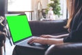 Woman using and typing on laptop with blank green desktop screen and coffee cups on table in modern loft cafe