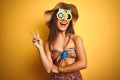 Woman on vacation wearing bikini and pineapple sunglasses over isolated yellow background smiling with happy face winking at the Royalty Free Stock Photo