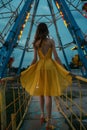 Woman in a vibrant yellow summer dress and matching shoes stands gracefully before a carousel at dusk, exuding charm and