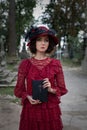 Woman in a vintage red dress and a black hat stands in a cemetery among the graves and holds a bible