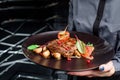 Woman waiter holding a dark round plate with grilled juicy steak with grilled mushrooms, tomatoes and cranberries. Royalty Free Stock Photo