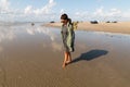 A woman walks barefoot through the water on Romo beach Denmark wearing a green coat and sunglasses with some cars on the sand in Royalty Free Stock Photo