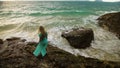 Woman walks on rock of sea reef stone, stormy cloudy ocean. Blue swimsuit dress tunic. Concept rest, tropical resort Royalty Free Stock Photo