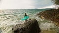 Woman walks on rock of sea reef stone, stormy cloudy ocean. Blue swimsuit dress tunic. Concept rest, tropical resort Royalty Free Stock Photo