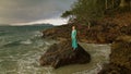 Woman walks on rock of sea reef stone, stormy cloudy ocean. Blue swimsuit dress tunic. Concept rest, tropical resort Royalty Free Stock Photo