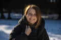 A woman in a warm black hooded down jacket rejoices at the good weather in a snowy park on a bright sunny winter day