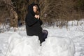 A woman in a warm long black down jacket with a hood is resting in a snowy park and rejoices in the bright sun and good weather