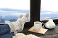 Woman in warm socks with hot drink enjoying view of winter mountain landscape from window Royalty Free Stock Photo