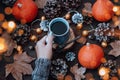 Woman in warm sweater holding cup of hot coffee with pumpkins, dry leaves and cones in background, autumn lifestyle, top view