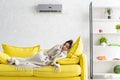 Woman warming with blanket while lying on yellow sofa under air conditioner at home