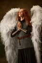 Woman-warrior with white wings and chainmail armor clasps hands in prayerful pose against vintage studio background.