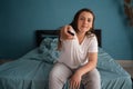 Woman watches TV sitting on the bed, using tv remote control in her hands. Morning or evening in the bedroom Royalty Free Stock Photo