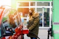 Woman wear medical mask at self-service gas station, hold fuel nozzle, refuel the car with petrol during corona virus Royalty Free Stock Photo