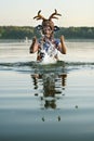 Woman wearing an Antlers Deer felted Hat and blue dress is staying waist-deep in the lake rising her hands up splashing the water