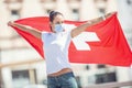 Woman wearing a face mask stands with arms wide open outdoors, holding a Swiss flag behind her