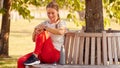 Woman Wearing Fitness Clothing Sitting On Seat Under Tree Checking Activity Monitor On Smartwatch