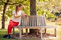 Woman Wearing Fitness Clothing Sitting On Seat Under Tree Checking Activity Monitor On Smartwatch