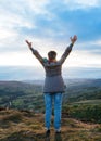 Woman wearing grey coat and jeans  raising her hands high and standing on top of mountain Royalty Free Stock Photo