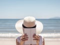 A woman wearing a hat and reading a book on the beach. Generative AI image. Royalty Free Stock Photo