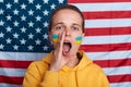 Woman wearing hoodie with flag of Ukraine on her cheek, yelling, screaming, posing isolated over flag of united states of america