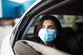 A woman wearing a medical sterile mask in a taxi car on a backseat looking sideway out of open window. Girl passenger waiting in a