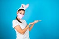 Woman wearing pink medical mask and rabbit ears  raises palms as if showing something  gestures over blank space. Coronavirus Royalty Free Stock Photo