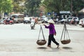 Woman wearing purple cloth and conical hat. Carry baskets with stuff inside with a carrying pole on the street in Hanoi, Vietnam