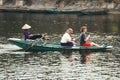 Woman wearing purple long arm t-shirt and conical hat rowing boat by her feet with tourists on the boat on the river at Trang An.