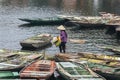 Woman wearing purple shirt, conical hat carry paddles and mat, walking on boat with many boats stop over the river at Trang An Gro
