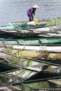 Woman wearing purple shirt, conical hat carry paddles and mat, walking on boat with many boats stop over the river at Trang An Gro