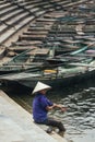 Woman wearing purple shirt, conical hat carry wash her feet in the river with empty rowing boats in the background at Trang An