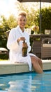 Woman Wearing Robe Sitting Outdoors Sitting With Feet In Swimming Pool Opening Champagne On Spa Day Royalty Free Stock Photo