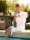 Woman Wearing Robe Sitting Outdoors Sitting With Feet In Swimming Pool Opening Champagne On Spa Day Royalty Free Stock Photo