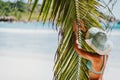 A woman wearing sun hat near coconut palm tree leaves on a tropical Anse Cocos sandy beach. La Digue, Seychelles