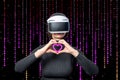 A woman wearing a virtual reality helmet shows with her hands the shape of a heart on a purple background of a matrix