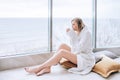 Woman in a white bathrobe enjoying sea view next to big window with cup of coffee. Panoramic window. Floor covering, Vacation Royalty Free Stock Photo