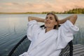 woman in white bathrobe relaxing in chair on lake footbridge at warm summer evening. nature enjoyment