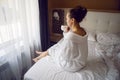 woman in a white bathrobe is sitting on the edge of the bed and drinking tea from mug at a large window in the hotel Royalty Free Stock Photo