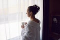 woman in a white bathrobe is sitting on the edge of the bed and drinking tea from mug at a large window in the hotel Royalty Free Stock Photo