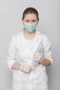 A woman in a white coat, mask and gloves holds a huge syringe in her hands.