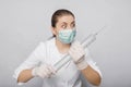 A woman in a white coat, mask and gloves holds a huge syringe in her hands.