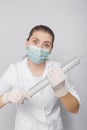 A woman in a white coat, mask and gloves holds a huge syringe in her hands.