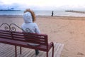 Woman in a white down jacket sitting on a bench on the beach and look into the distance. Sea winter, back view. Royalty Free Stock Photo