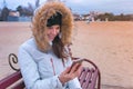 Woman in a white down jacket sitting on a bench on the beach and looking amazing video at mobile phone. Royalty Free Stock Photo