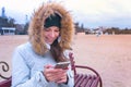 Woman in a white down jacket sitting on a bench on the beach and looking amazing video at mobile phone. Royalty Free Stock Photo