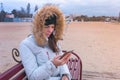 Woman in a white down jacket sitting on a bench on the beach and looking sad video at mobile phone. Royalty Free Stock Photo