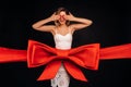 A woman in a white dress as a gift covers her eyes with her hands on a black background wrapped in a festive ribbon Royalty Free Stock Photo