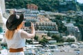 A woman in a white dress, and a hat stands on the beach in Positano and makes selfie photo on smartphone. Travel and vacation