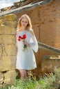 Woman in white dress standing near stone wall and holding poppies plants